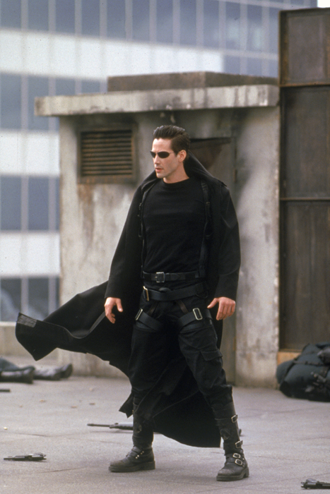 Full shot of Keanu Reeves as Thomas A. Anderson/Neo wearing sunglasses and coat/duster standing on skyscraper rooftop.