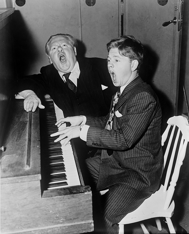 Medium shot of Mickey Rooney sitting at and playing piano with his father, character actor Joe Yule. Both are singing back stage of an MGM set, March 1940.